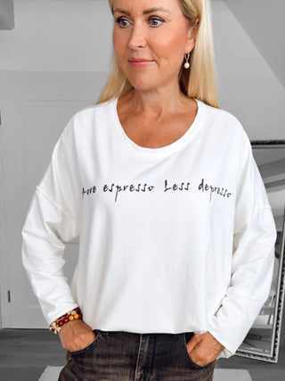Frau trägt OFF#DLY Shirt Espresso white, Oversize-Schnitt mit „more espresso less depresso“-Schriftzug. Kombiniert mit dunklen Jeans und warmen Armbändern. Ideal für Kaffee-Liebhaber.