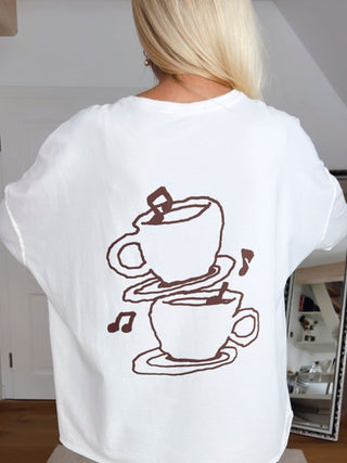 OFF#DLY Shirt Espresso white zeigt auf der Rückseite zwei handgezeichnete Kaffeetassen, übereinander gestapelt, mit fallendem Zuckerwürfel und Noten, getragen von jemandem mit langen blonden Haaren.