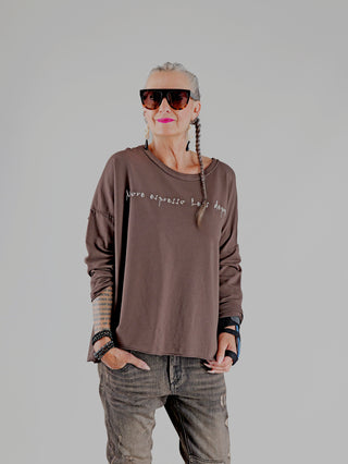 Frau in OFF#DLY Shirt Espresso brown mit weißem Schriftzug, oversized und aus Baumwollmix. Kombiniert mit grauen Jeans, großen Sonnenbrillen, Haarzöpfen und auffälligen Accessoires. Perfekt für kaffeeliebende Trendsetter.