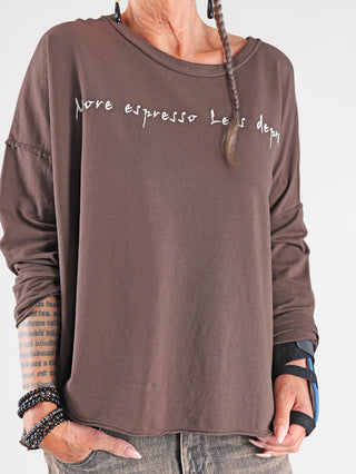 Person trägt OFF#DLY Shirt Espresso brown mit lockerem Schnitt, Slogan More espresso, less depresso auf der Brust, schwarze Teardrop-Ohrringe, Text-Tattoos am Arm und blauen Handgelenkstütze.