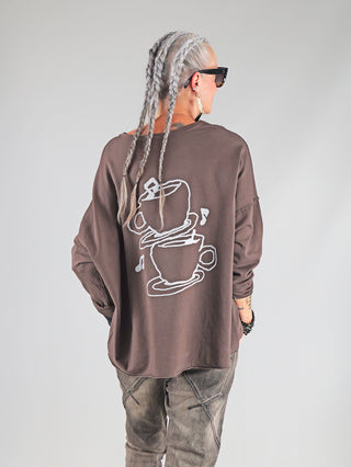 Person von hinten in OFF#DLY Shirt Espresso brown mit weißem Kaffeetassen-Print, langen silbernen Haaren, schwarzen Sonnenbrillen und graubraunen Jeans. Das Shirt hat einen oversize Schnitt.