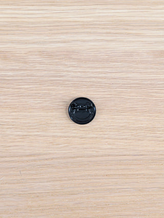 Rückseite eines runden, schwarzen Kunststoff-Ansteckbuttons mit Nadelverschluss, auf Holzoberfläche liegend. Produkt: OFF#DLY Sticker Smiley.