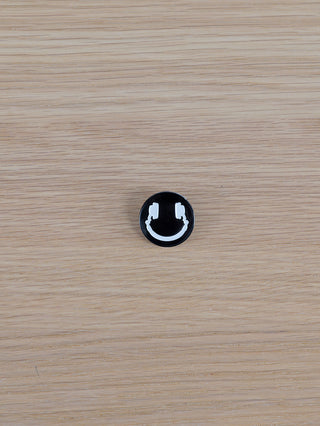 Ein runder OFF#DLY Sticker Smiley Ansteckpatch, zeigt ein lächelndes Gesicht mit Kopfhörern, liegt auf einer hellen Holzoberfläche.