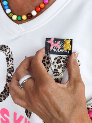 Hand hält das OFF#DLY Sticker Animal Ansteckpatch mit Nadel auf weißem Stoff, verziert mit pinkem Ballonhund und gelbem Teddybär, sowie silbernen Ketten. Träger trägt Leopardenprint-Shirt und bunte Halskette.