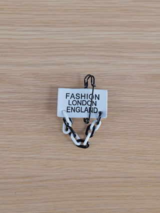 OFF#DLY Sticker Fashion: Weißes Stoffetikett mit FASHION LONDON ENGLAND, befestigt an einer übergroßen schwarzen Sicherheitsnadel, hängt an einer kurzen, abwechselnd schwarzen und weißen Kunststoffkette auf hellem Holz.