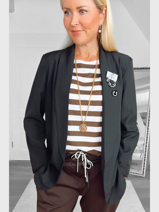 Frau trägt einen schwarzen Blazer mit OFF#DLY Sticker Fashion Ansteckpatches auf dem linken Revers, kombiniert mit gestreiftem Oberteil und brauner Hose, posierend mit Händen in den Taschen vor einem Spiegel.