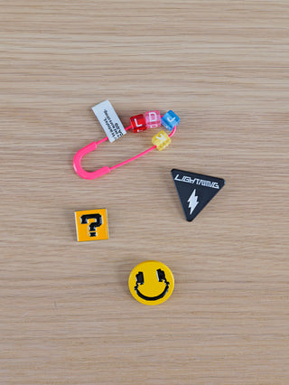 OFF#DLY Sticker Smiley Set bestehend aus einem pinken Sicherheitsclip mit Buchstabenwürfeln, einer schwarzen Dreiecksnadel mit LIGHTNING, sowie zwei gelben Ansteckern mit Fragezeichen und Smiley.