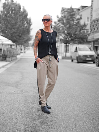Person trägt OFF#DLY Pant Turin taupe mit schwarzem Seitenstreifen, kombiniert mit einem schwarzen, ärmellosen Oberteil und schwarzen Stiefeletten. Accessoires: große Ohrringe, Halskette, Armbänder, und Sonnenbrille.