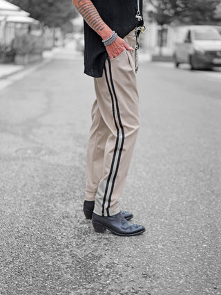 OFF#DLY Pant Turin taupe: Person trägt leicht konische, hellbeige Hose mit schwarzen Streifen an den Nähten, kombiniert mit schwarzen Stiefeletten und silbernen Armbändern, auf einer gepflasterten Straße.