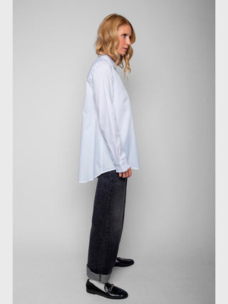 Frau in lockerem, langärmligem Emily Bluse 4890-157120 white, das über die Hüften fällt, mit Hemdblusenkragen und Rüschenblende, kombiniert mit dunklen, gerade geschnittenen Jeans und schwarzen Loafern.