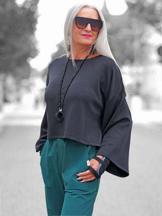 Ältere Frau trägt den BSB Pulli black mit weiten Ärmeln und lockerem Schnitt, kombiniert mit teal-grünen Hosen. Der Pulli betont urbanen Chic und Komfort.