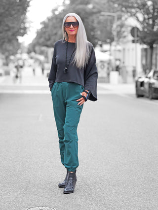 Frau trägt Penn&Ink Trousers forest, stylische Jogpant in Teal-Grün mit elastischem Bund und sportiven Rippdetails, kombiniert mit schwarzer Bluse und Stiefeletten auf städtischer Straße.
