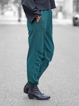 Penn&Ink Trousers forest: Teal-grüne Joggerhose im Cargo-Stil mit elastischen Bündchen, getragen mit schwarzen Lederstiefeletten. Eine Hand in der Tasche, sichtbare Armbänder am Handgelenk.