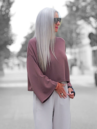 Eine Person trägt den BSB Pulli chocolate mit weiten Ärmeln, kombiniert mit losen grauen Hosen. Der Pulli zeigt urbanen Chic, unterstützt durch Accessoires wie Ringe und ein Armband.