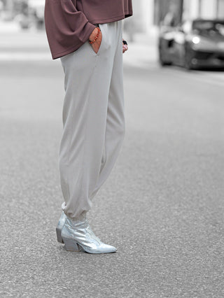 Person trägt Penn&Ink Trousers T 1182 sand, locker sitzende, knöchelcinchende Jogpants mit bequemer Passform, kombiniert mit glänzenden, spitzen Cowboy-Boots. Hand in Tasche, Armband sichtbar.