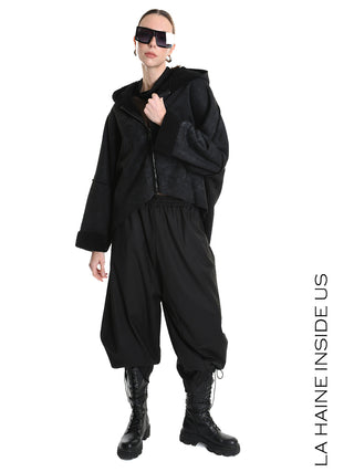 Eine Person trägt die La Haine Jacket black W5168, eine übergroße schwarze Jacke mit Reißverschluss. Kombiniert mit weiten Jogginghosen, robusten Kampfstiefeln, großen Sonnenbrillen und einfachen Creolen.