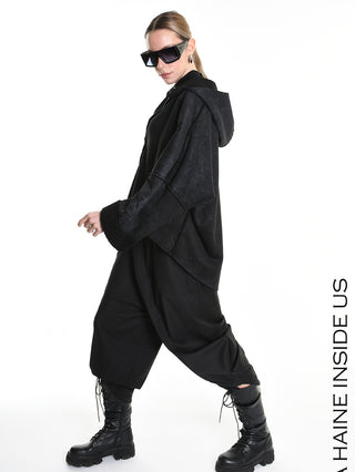 Person mit schwarzer, oversized La Haine Jacket black W5168 mit weiten Ärmeln, kombiniert mit baggy Haremshosen und klobigen Kampfstiefeln. Trägt übergroße Sonnenbrille, Haare im tiefen Pferdeschwanz.