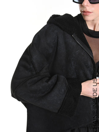 La Haine Jacket black W5168: Schwarze sherpa-gefütterte Kapuzenjacke mit sichtbarem Reißverschluss und geräumigen Ärmeln, darunter ein schwarzes, durchsichtiges Top. Teilweise sichtbare Hand und übergroße dunkle Sonnenbrille.