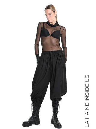 Das Model trägt die La Haine Pant black LW5164, eine verkürzte, schwarze Haremhose mit elastischem Kordelzugbund und Seitentaschen, kombiniert mit hohen Schnürstiefeln für einen urbanen Look.