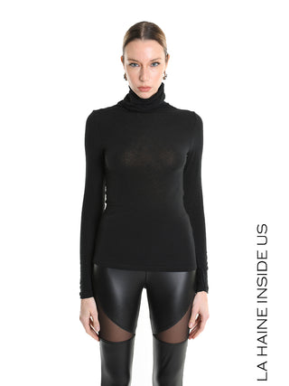 Frau in schwarzem, figurbetontem La Haine Pulli LW5232 mit Rollkragen, kombiniert mit hochtaillierten Leggings mit Lederoptik und Mesh-Einsätzen, vor weißem Hintergrund.