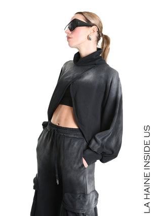 Junge Frau trägt La Haine Sweater LW5226 black mit überkreuztem Design und cowl-neck. Kombiniert mit Cargo-Jogpants und schwarzen Sonnenbrillen, präsentiert sie einen urbanen, futuristischen Look.