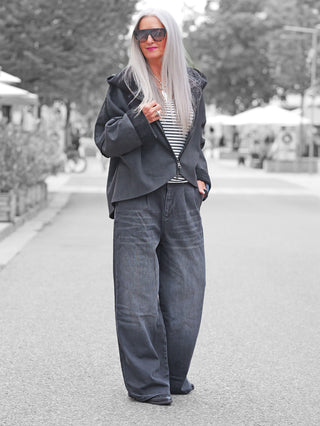Frau in Stadt trägt Gitta Banko Rylee W.Black, weite, hochgeschnittene Jeans mit Bundfalten, kombiniert mit schwarzer Jacke und gestreiftem Top, betont durch geometrische Sonnenbrille und silberne Accessoires.