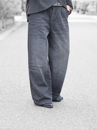 Person trägt Gitta Banko Rylee W. Black Jeans, weit geschnitten, verdeckt größtenteils schwarze Schuhe, kombiniert mit einem schwarzen Jackenrand und einem gestreiften Shirt. Hand lässig in der Tasche.