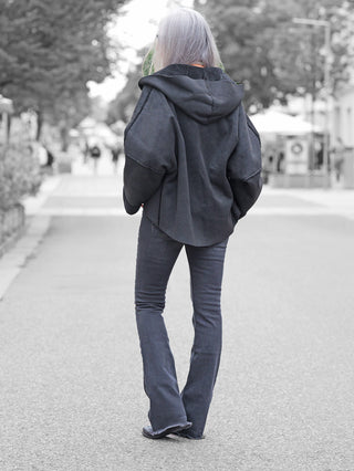 Person in schwarzer Kapuzenjacke trägt Gitta Banko Alba Flared Jeans W. Black. Die schmal geschnittene Jeans mit ausgestelltem Bein kombiniert sich mit lässigem Stil auf einer belebten Fußgängerstraße.
