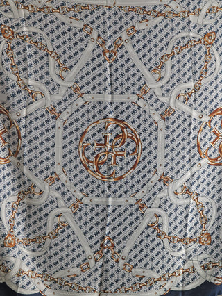 GUESS Noelle Scarf 130x130 BLU mit navyblauen Emblemen auf weißem Hintergrund, goldenen Kettengürtelmotiven und zentralem Medaillon, aus 100% Polyester, quadratisch, elegant und dekorativ.