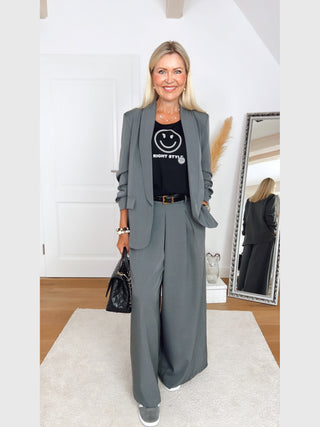 Frau trägt OFF#DLY Hose Giulia Grau, weite, elegante Hose, kombiniert mit schwarzem T-Shirt, Gürtel und Sneakers. Outfit reflektiert lässigen Chic und feminine Raffinesse in modernem Raum.