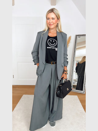 Die stilvolle Frau trägt den OFF#DLY Blazer Eleonora Grau, locker geschnitten mit gerafften 3/4-Ärmeln, kombiniert mit einem Statement-Shirt und weiten grauen Hosen für einen modernen Look.