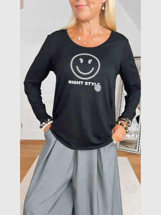 Doris Streich Shirt 268270 schwarz mit Strass-Smileymotiv und RIGHT STYLE-Schriftzug, kombiniert mit hellgrauen, weiten Hosen. Accessoires: Armbänder, Armbanduhr, zarte Halskette und lange Ohrringe.