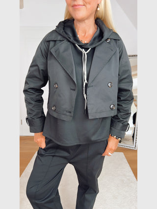 Die OFF#DLY Jacke Alessia black zeigt sich als schwarze, kurz geschnittene, doppelt geknöpfte Jacke mit Reverskragen, über einem Hoodie getragen, kombiniert mit eleganten Accessoires.