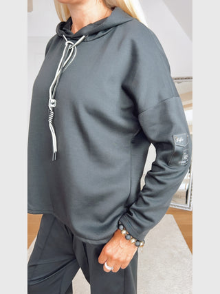 Person trägt das OFF#DLY Sweatshirt Life black, einen lockeren, dunklen Hoodie mit weißen Kordeln und Patch „Life“ am Ärmel. Dazu passende Hose und auffälliger Schmuck am Handgelenk.