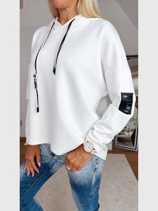 Person trägt OFF#DLY Sweatshirt Life white, einen weißen Hoodie mit schwarzen Kordeln und Patch am Ärmel, kombiniert mit blauen Jeans, goldener Uhr und Ring.