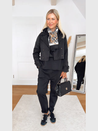 Die OFF#DLY Jacke Alessia black, eine kurze Trenchcoatjacke mit Reverskragen und doppelter Knopfleiste, wird von einer Frau in einem lichtdurchfluteten Raum getragen.