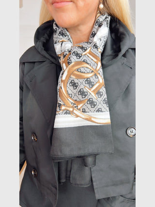 GUESS Noelle Scarf 130x130 BLA, drapiert um den Hals, zeigt graue, schwarze und weiße Töne mit 4G-Logo und goldfarbenen Kettengürtel-Print, getragen über einem schwarzen Trenchcoat.