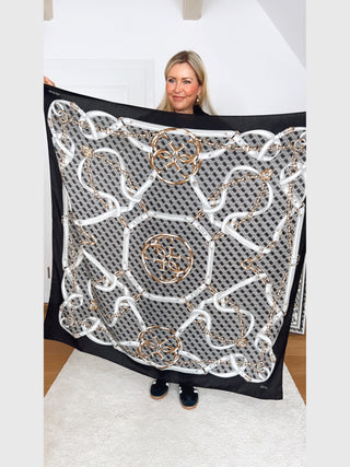 GUESS Noelle Scarf 130x130 BLA: Person hält quadratisches Tuch mit schwarzem Rand, weißen und goldenen Kettengürtel-Motiven in achteckigen Rahmen und kleinem GUESS-Logo in einer Ecke.