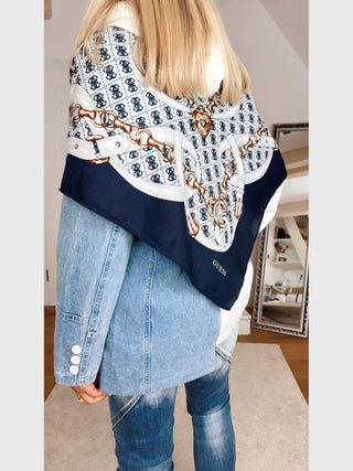 Person von hinten mit GUESS Noelle Scarf 130x130 BLU über den Schultern, zeigt Kettengürtel-Print und 4G-Logo, getragen über einer hellen Jeansjacke, kombiniert mit dunkleren Jeans mit Knie-Details.