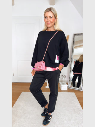 Frau trägt OFF#DLY Joggpant Riva black mit Gürtel, die durch ihren modernen, leicht glänzenden Schnitt und sportliche Aufschläge besticht, kombiniert mit Adidas-Sneakern und einer pinken Crossbody-Tasche.