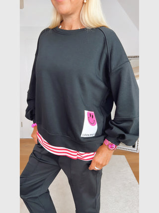 Frau trägt OFF#DLY Sweat Smile black: schwarzer, locker geschnittener Pullover mit pinkem Smiley-Patch LOOK BACK, kombiniert mit rot-weiß gestreiftem Shirt und schwarzen, konischen Hosen.