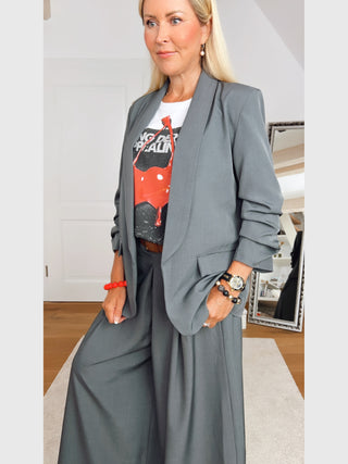 Person trägt OFF#DLY Blazer Eleonora Grau mit gerafften Ärmeln, über einem weißen T-Shirt mit rotem Design. Kombiniert mit Schmuck, steht sie vor einem Spiegel auf einem hellen Teppich.