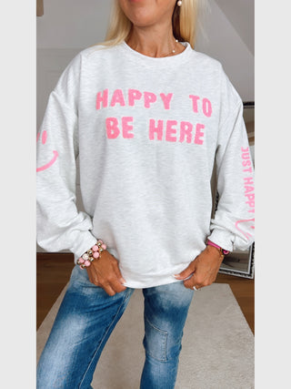 Person trägt den OFF#DLY Sweater HAPPY grey melange mit pinkem Schriftzug „HAPPY TO BE HERE“ auf der Brust und „JUST HAPPY“ auf den Ärmeln, kombiniert mit blauen Jeans und Armbändern.