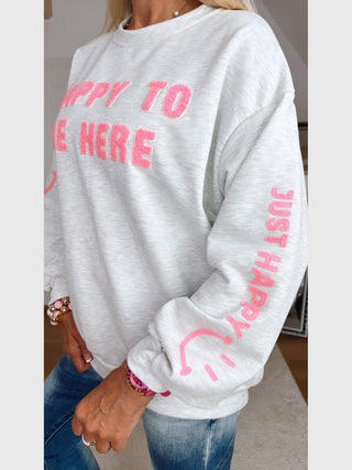OFF#DLY Sweater HAPPY grey melange: Person trägt grauen Pullover mit pinkem 3D-Text HAPPY TO BE HERE auf der Brust und JUST HAPPY mit Smiley auf dem Ärmel. Kombiniert mit blauer Jeans.