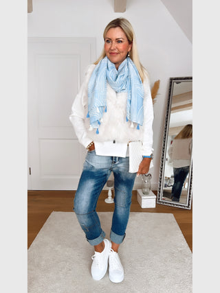 Person trägt den OFF#DLY Schal Waves Fransen blau/beige um den Hals, kombiniert mit blauer Jeans und weißen Sneakers, in einem hellen Raum mit Spiegel im Hintergrund.