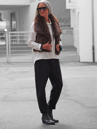 Person trägt die OFF#DLY Fellweste Martina brown aus weichem Kunstfell. Die Weste ergänzt den weißen Longsleeve und die schwarzen, locker geschnittenen Hosen für einen modernen, urbanen Look.