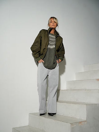 Junge Frau im Gitta Banko Pulli Genevieve army, einem locker geschnittenen Norwegerpullover mit cremefarbenem Muster, kombiniert mit hellgrauen Jeans und schwarzen Schuhen, lehnt lässig an einer Wand.