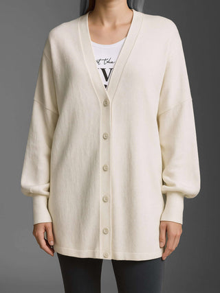 Die Cotton Candy Cardigan Malea offwhite/mela zeigt einen oversized Strickcardigan mit tiefem V-Ausschnitt, Ballonärmeln und gerippten Bündchen, getragen über einem T-Shirt, kombiniert mit dunklen Slim-Fit-Hosen.