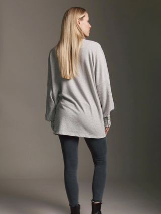 Eine Frau trägt den Cotton Candy Cardigan Malea grey/melan mit Ballonärmeln und Knopfmanschetten, kombiniert mit grauen Leggings und schwarzen Stiefeletten, von hinten gesehen.