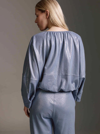 Frau von hinten in schimmerndem, blauen Set mit weitem Schnitt. Oberteil mit gerafftem Hals, weiten Schultern und langen Ärmeln, kombiniert mit weiten Hosen. Passend zur Cotton Candy Jacket Maaija blue ash.